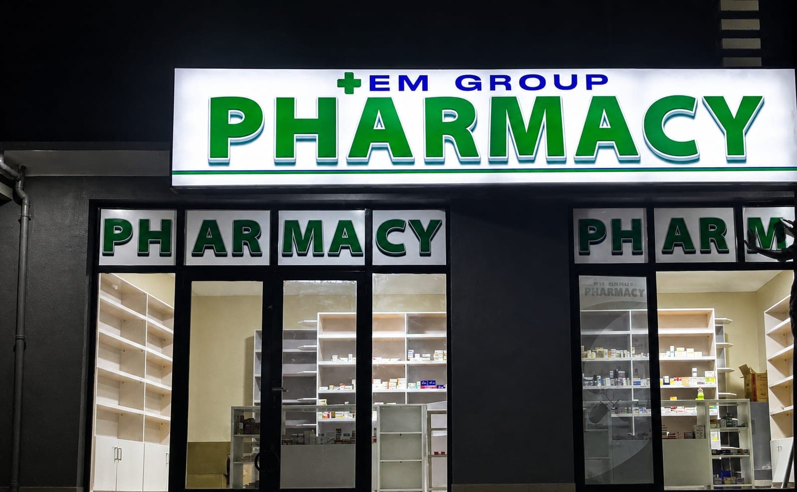 A pharmacy lit up after dark, its illuminated sign reading EM GROUP PHARMACY in green above wide windows, with dispensary shelving visible inside.