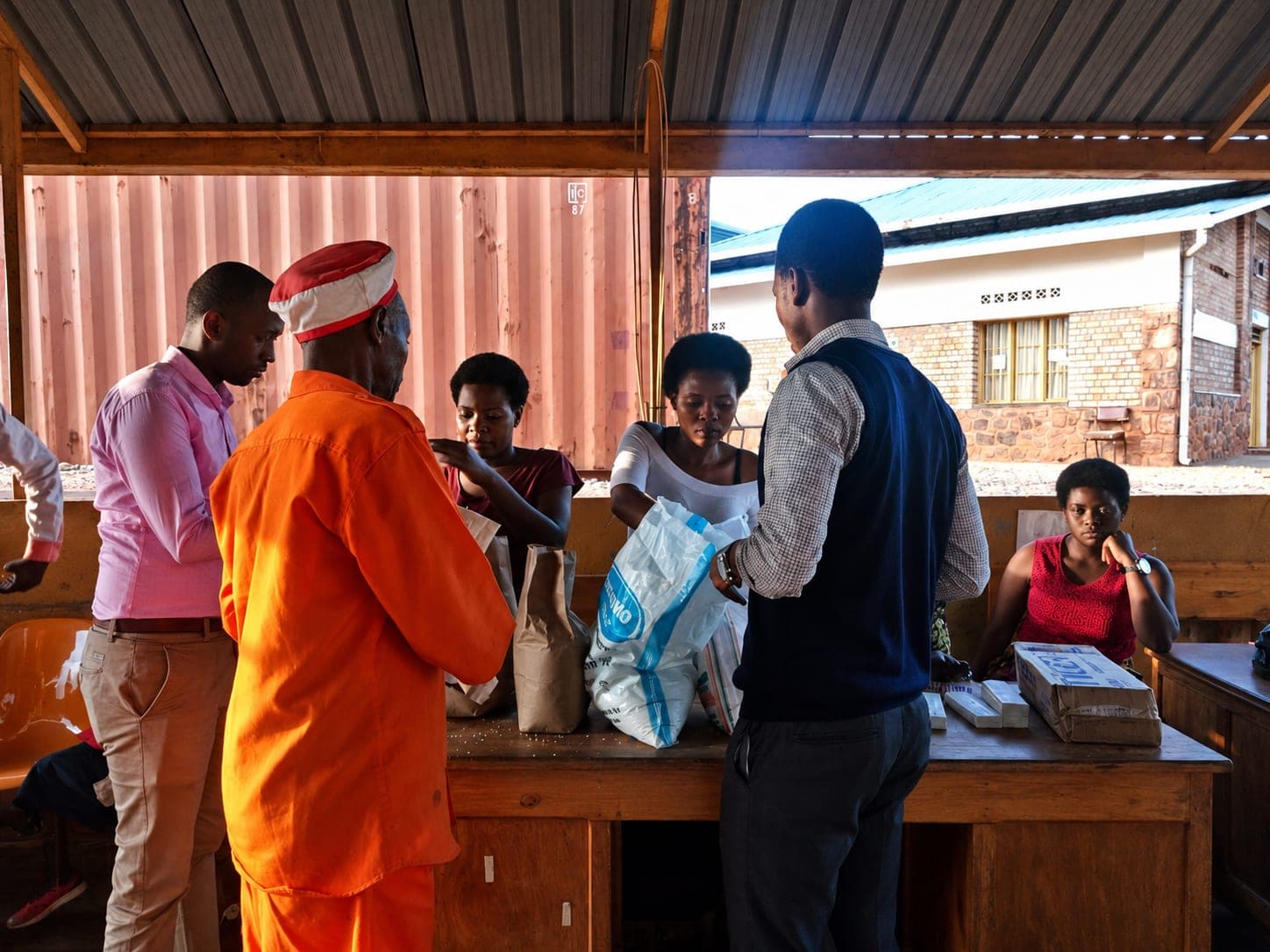 People gathered at a long wooden counter under a corrugated roof, weighing out and handing over paper bags and a sack of flour.